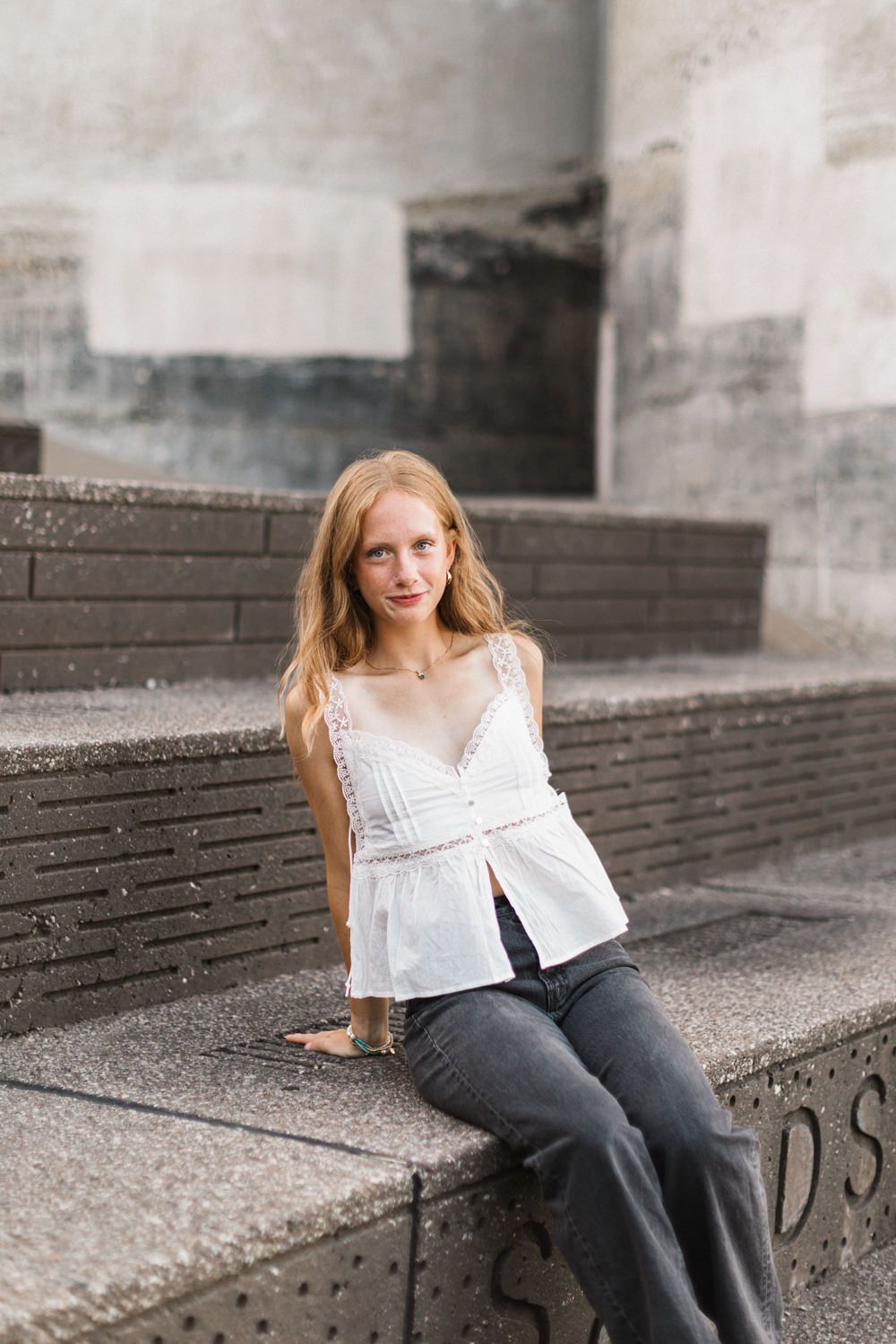 Person in white top and dark pants sits casually on concrete steps against an urban backdrop.