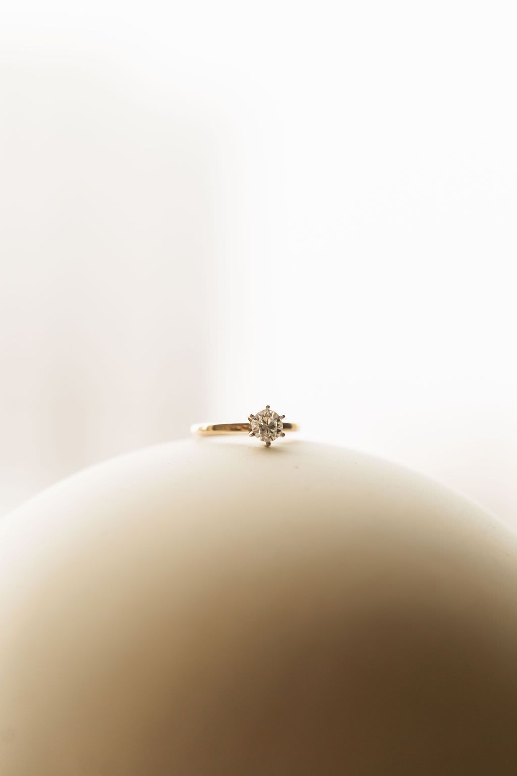 A delicate gold engagement ring with a diamond solitaire rests on a curved white surface in soft lighting.