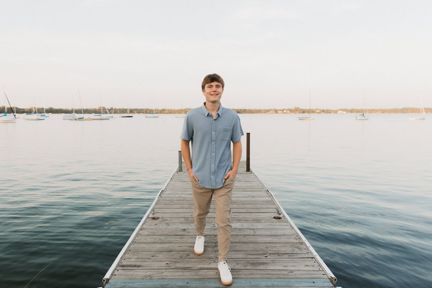 Young man in blue shirt standing on wooden dock overlooking calm lake at sunset.