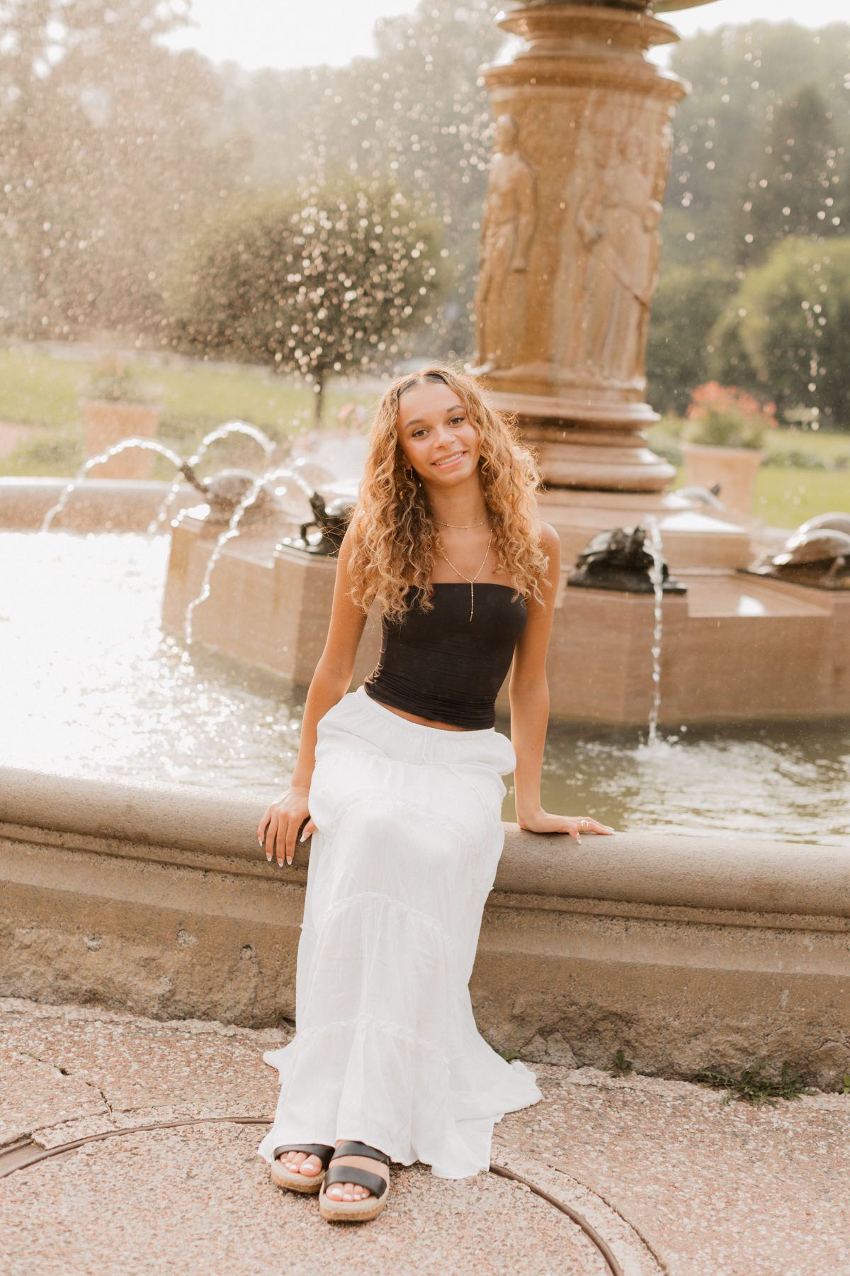 Young woman with curly hair sits on a fountain edge wearing a black crop top and white maxi skirt outdoors.