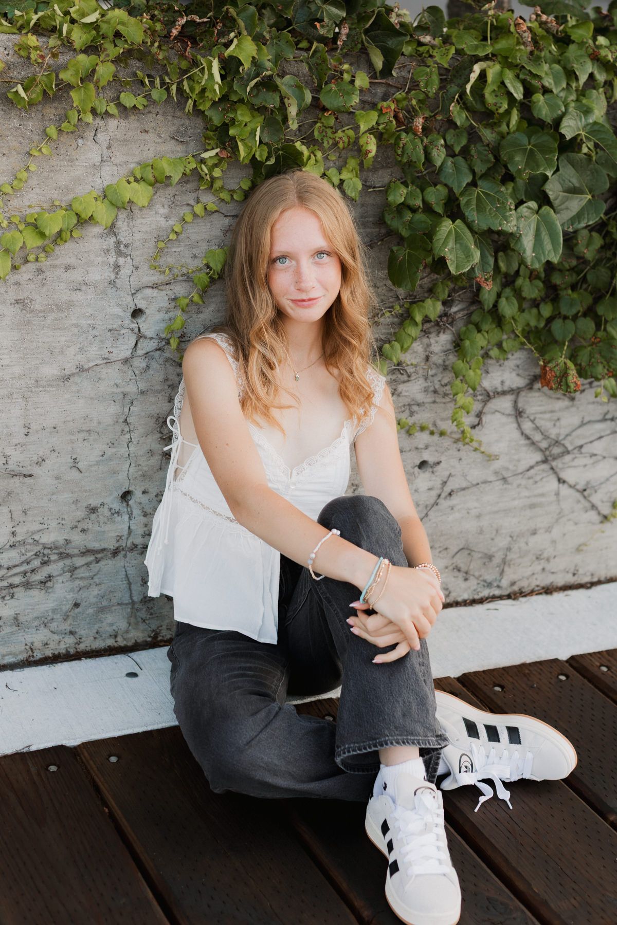 Young woman in white top and black jeans sitting on wood deck against ivy-covered concrete wall.