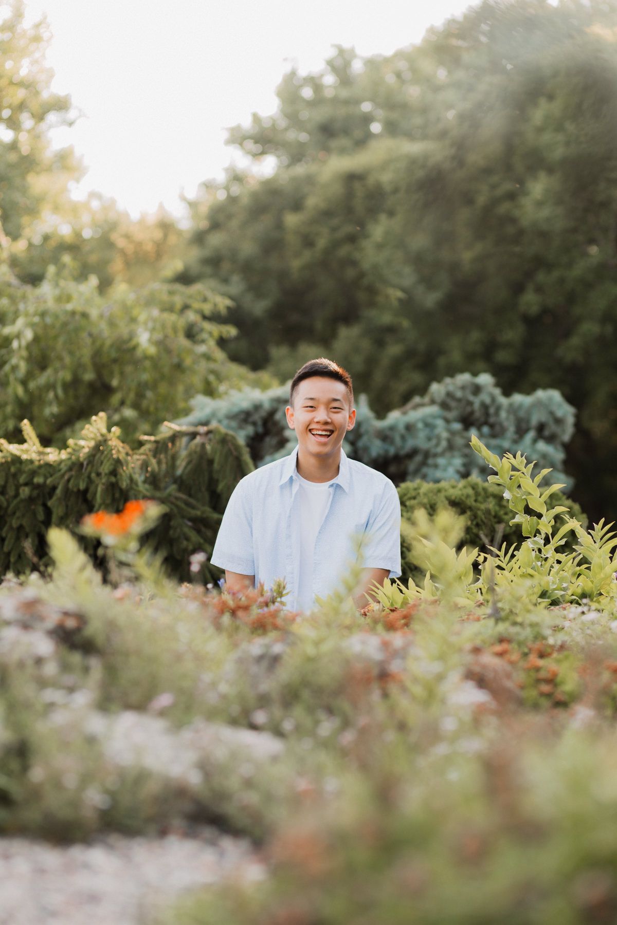 Smiling young Asian man in light blue shirt standing among colorful garden plants in warm sunlight.