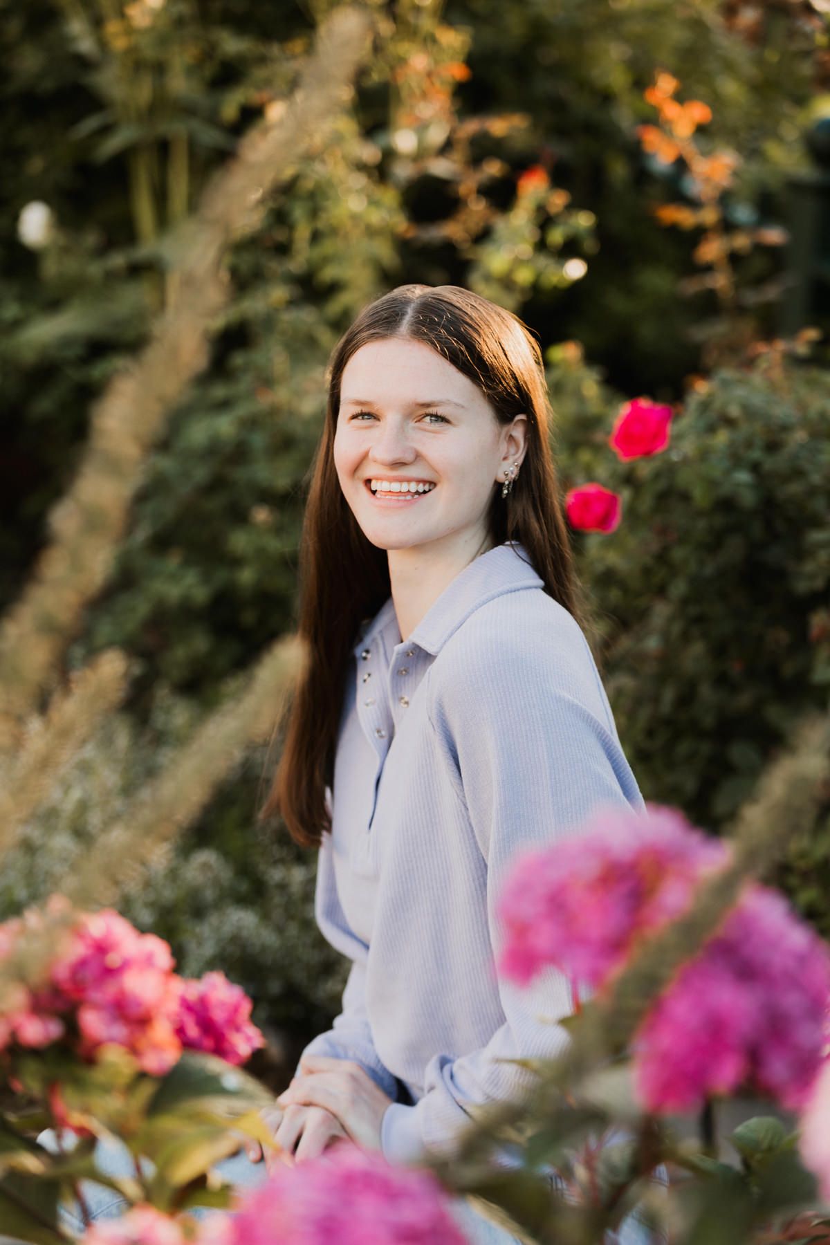 Smiling brunette teen girl in light blue shirt posing among colorful garden flowers at golden hour.