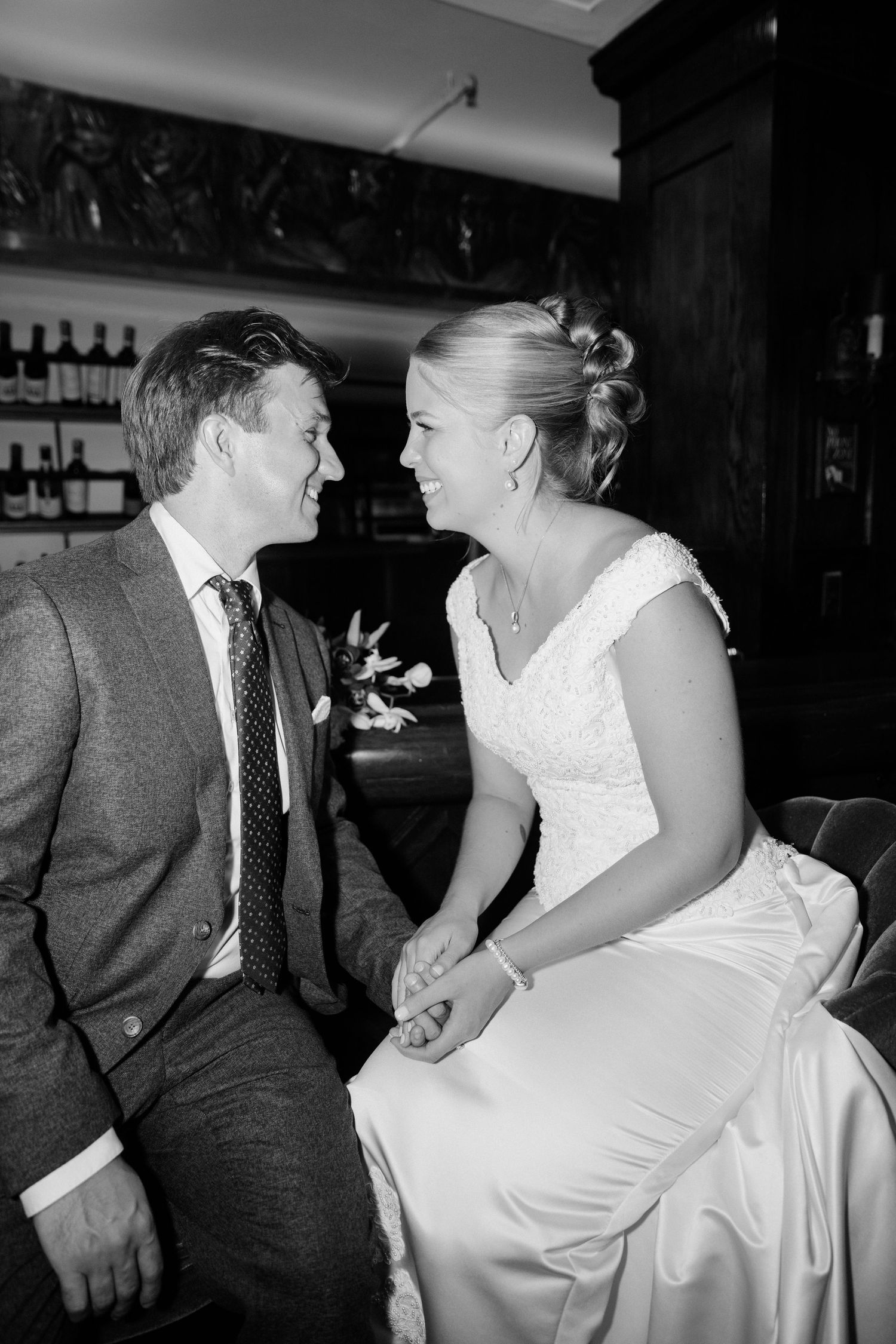 Black and white photo of bride and groom sitting close, smiling at each other in a warmly lit bar setting.