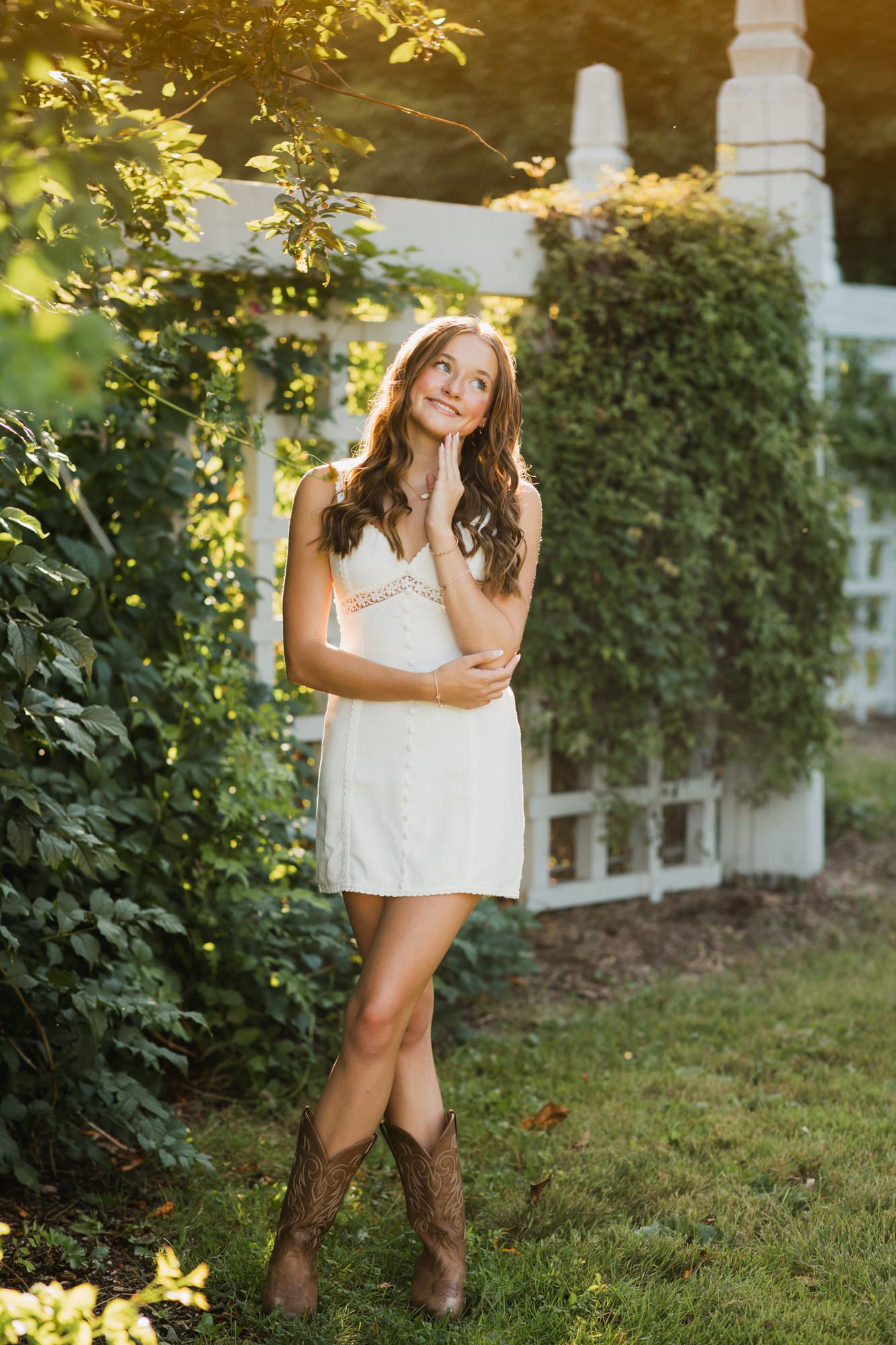 Young woman in white sundress and cowboy boots poses gracefully in a lush garden near a white fence.