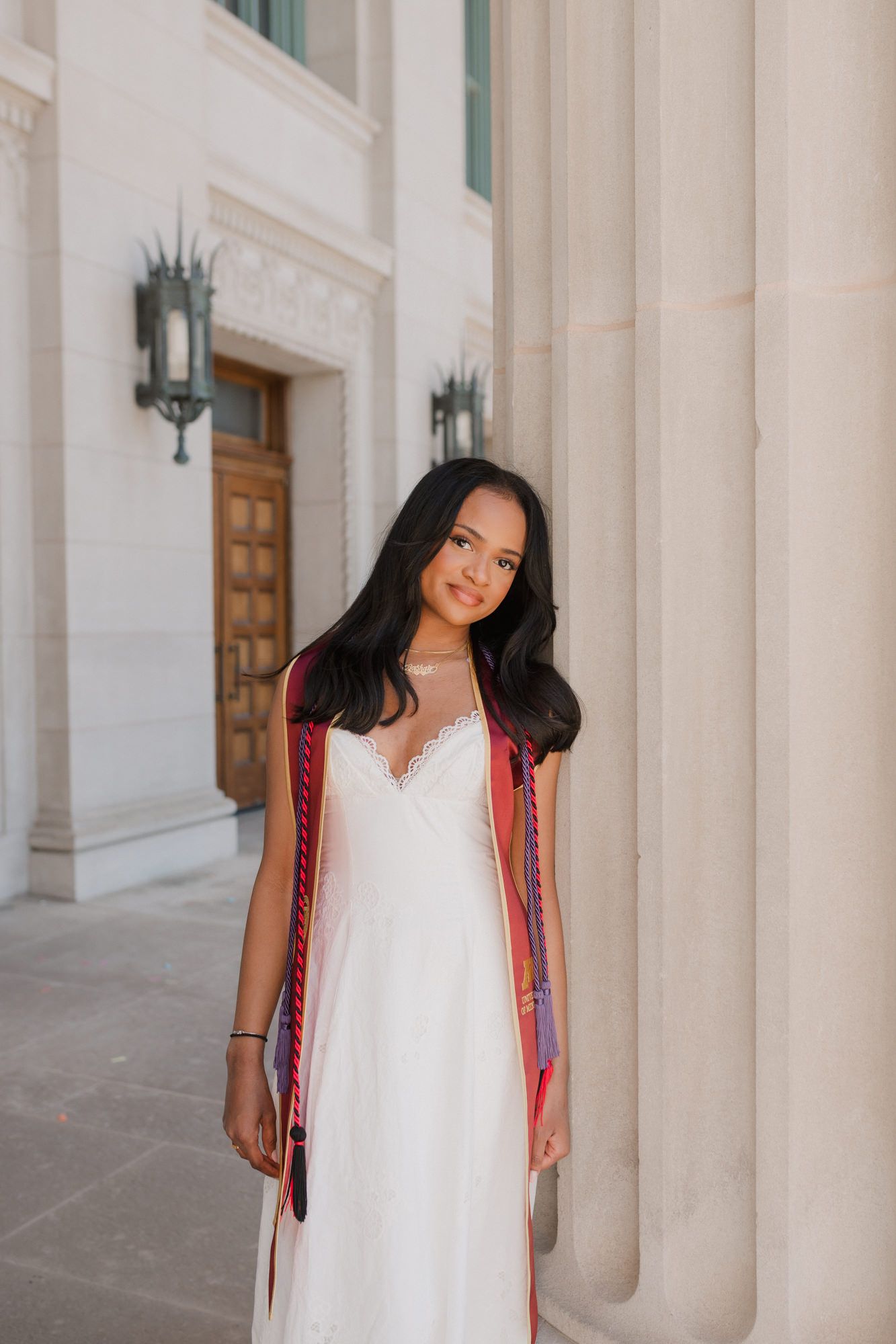 Young woman in white dress with colorful graduation stole poses beside large stone columns at university building.