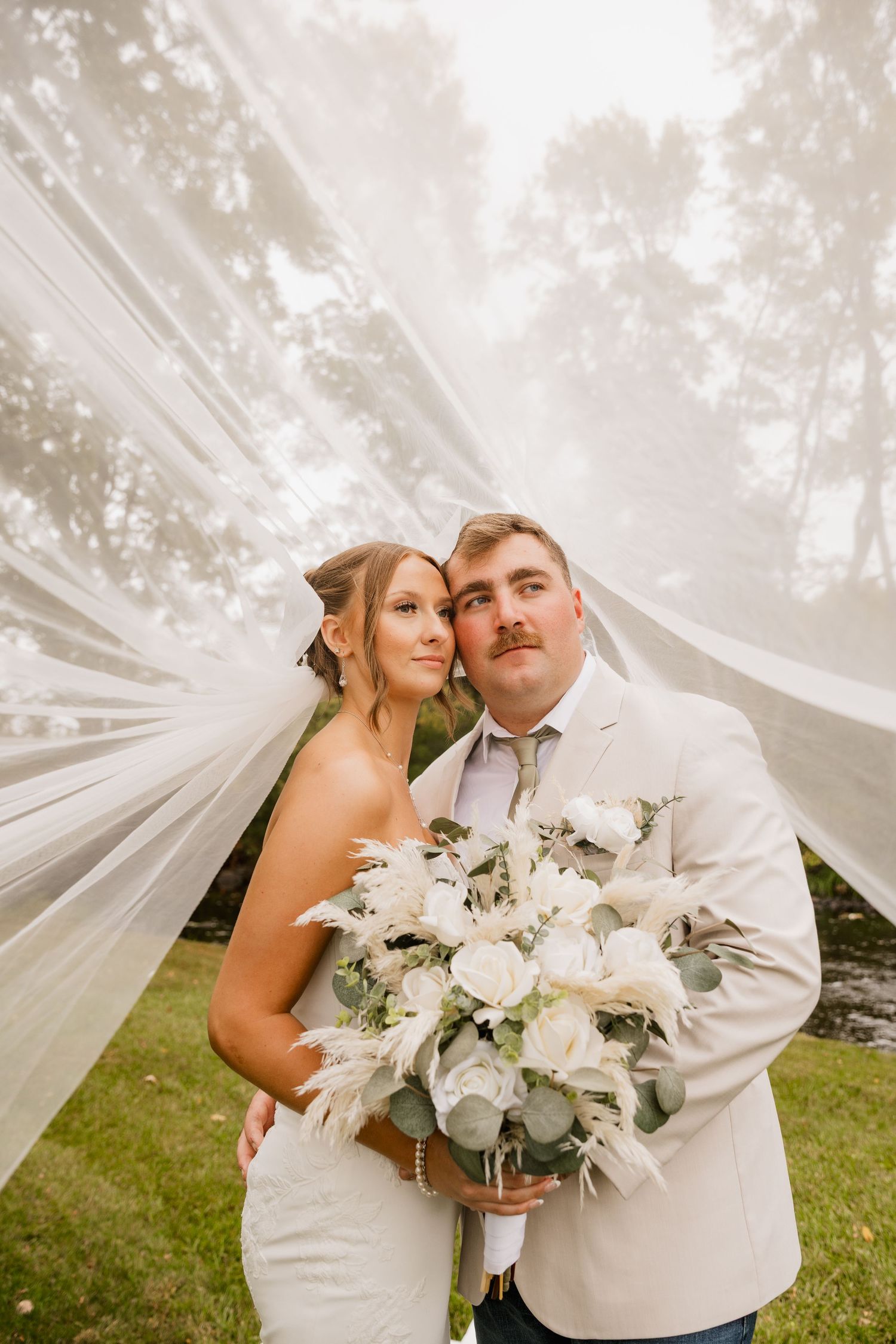 Bride and groom pose under flowing white veil outdoors, holding white rose bouquet with eucalyptus on their wedding day.