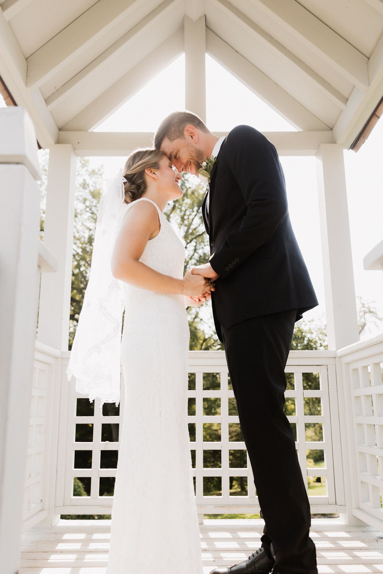 Bride and groom share a romantic moment holding hands in a white gazebo, dressed in wedding attire.