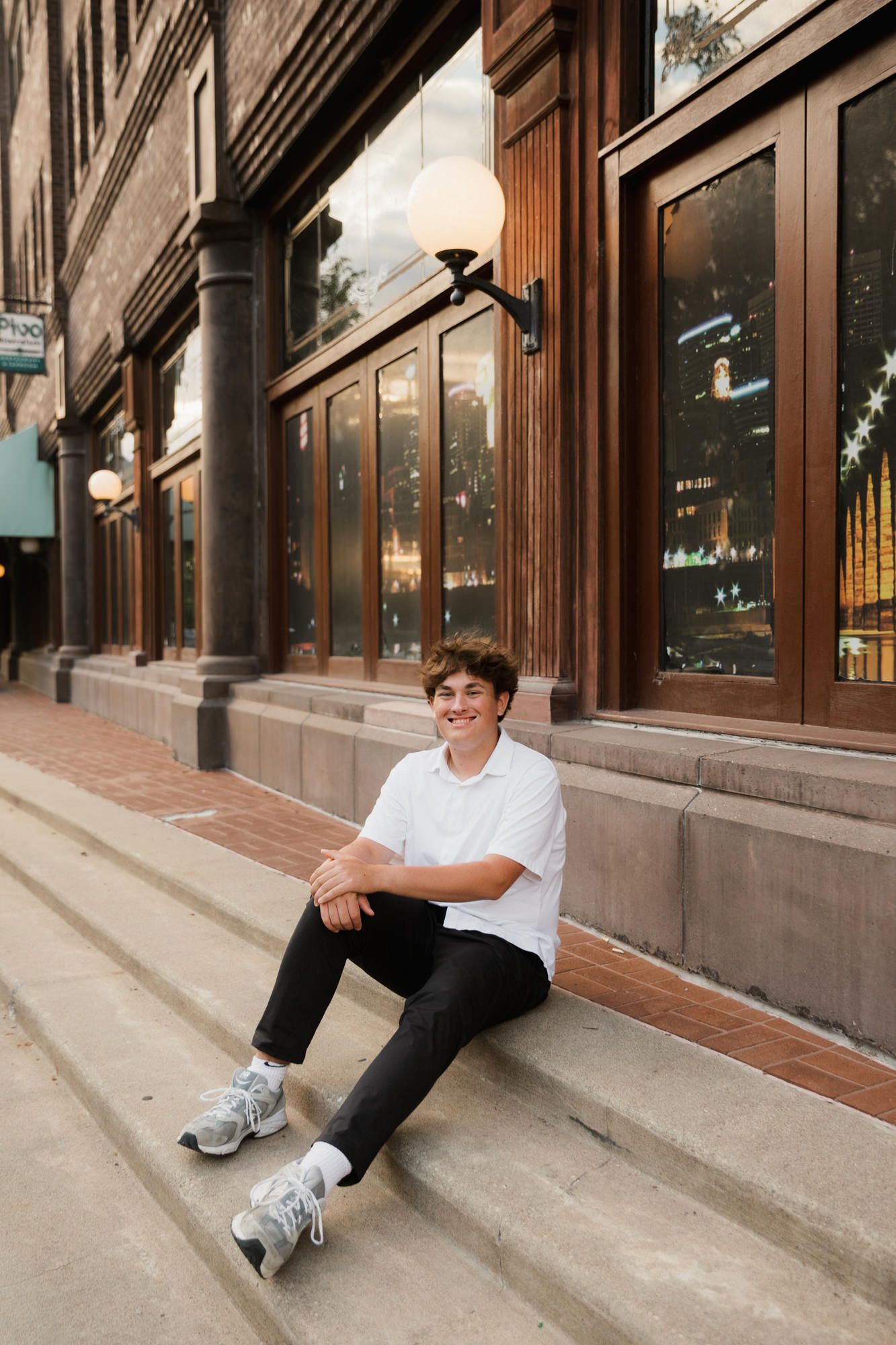 Young man in white tee and black pants sitting on urban steps, smiling confidently outside a brick building.