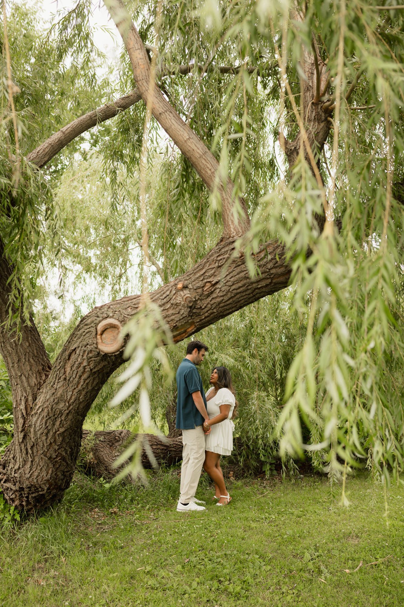 Couple standing romantically under a large willow tree surrounded by lush green foliage in an outdoor park setting.