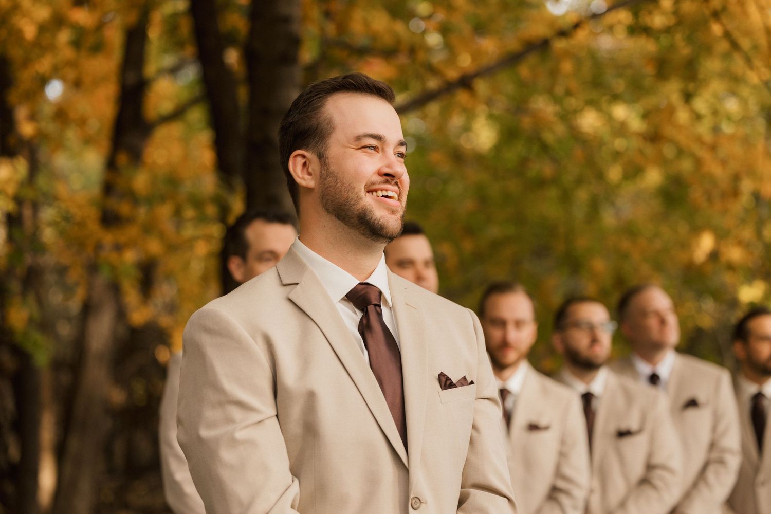 Smiling groom in a tan suit and burgundy tie stands with groomsmen amid golden autumn leaves at an outdoor ceremony.