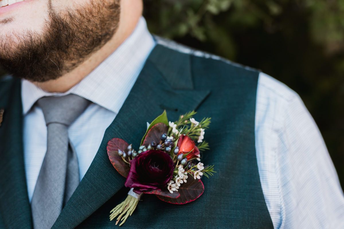 Close up detail shots of burgundy floral boutonnieres with berries pinned to grey suit lapels at a wedding.