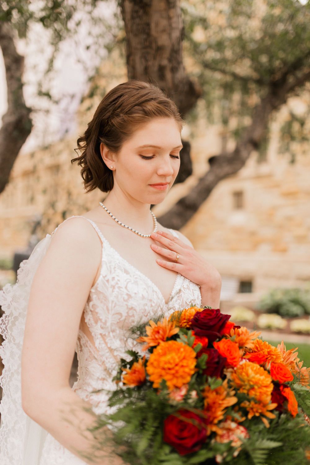 A bride in a lace wedding dress holds a vibrant autumn bouquet with orange and red flowers in a natural outdoor setting.