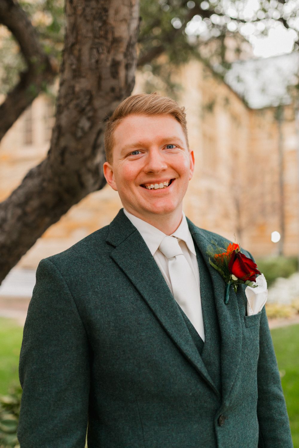 A groom in a dark green suit with a red rose boutonniere smiles outdoors against a blurred natural background.