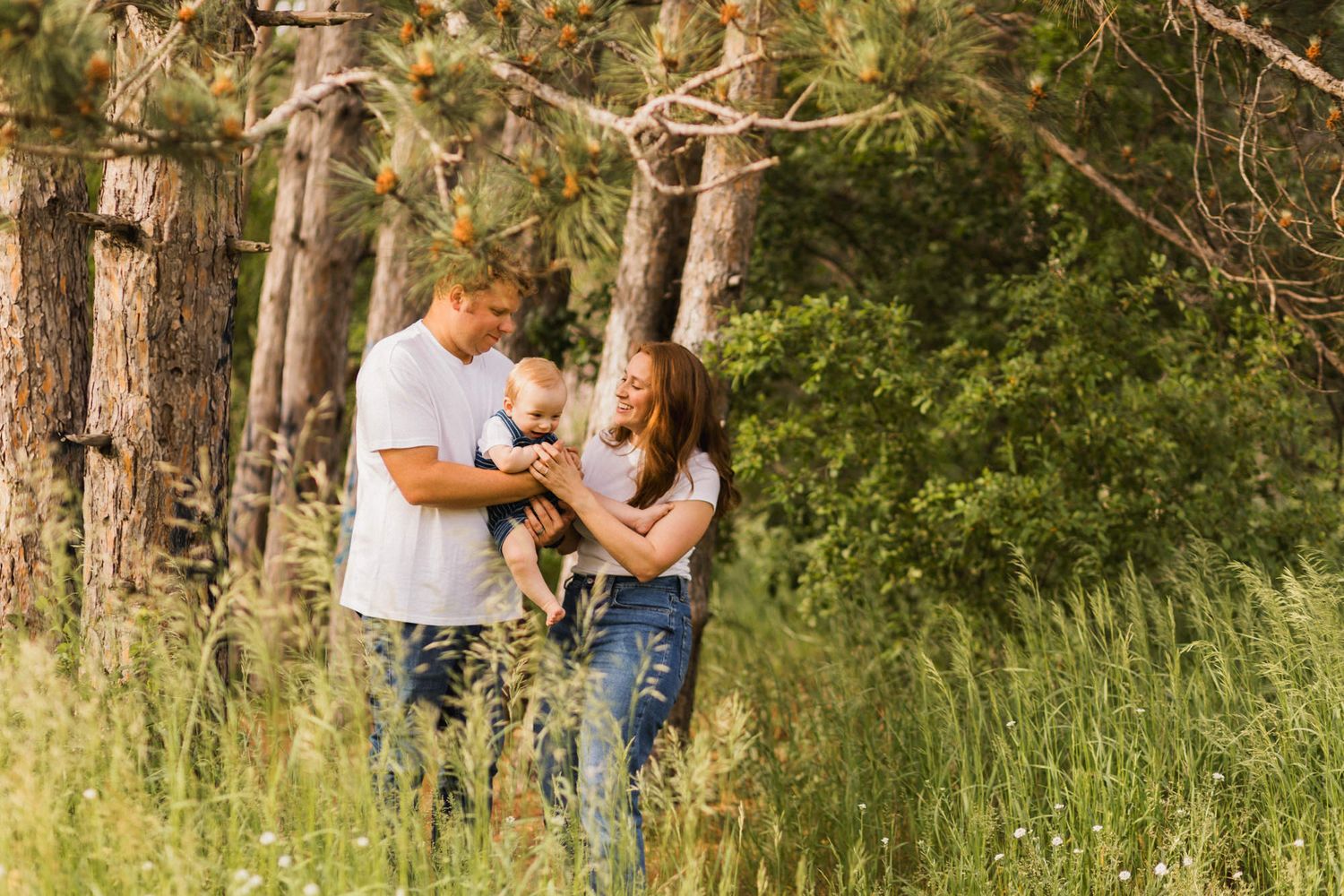 A couple stands in a lush green forest holding a small dog, enjoying a sunny outdoor family photo session.