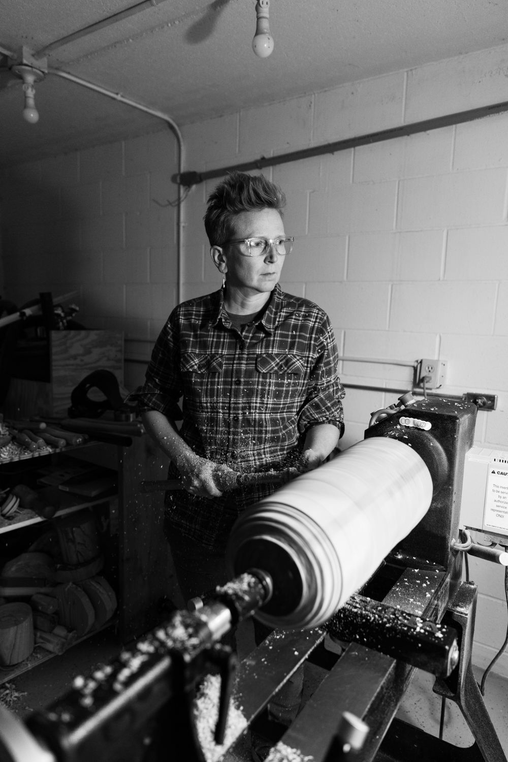 Woman in plaid shirt and glasses operates a large wood lathe in a workshop, with wood shavings scattered around.
