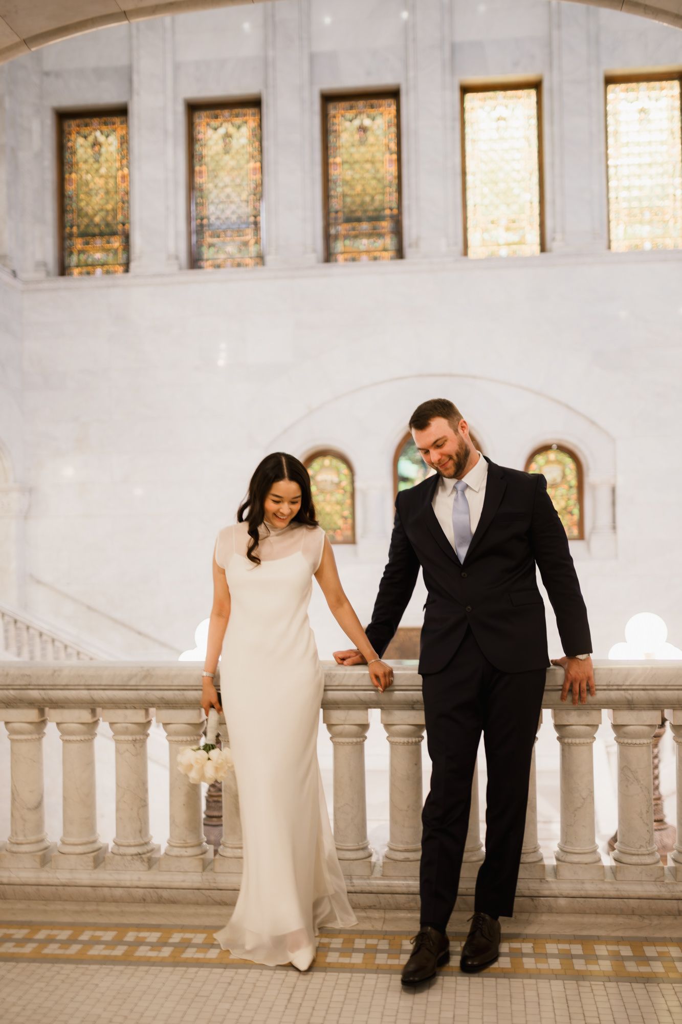 Bride and groom hold hands on marble balcony with stained glass windows in elegant wedding venue.