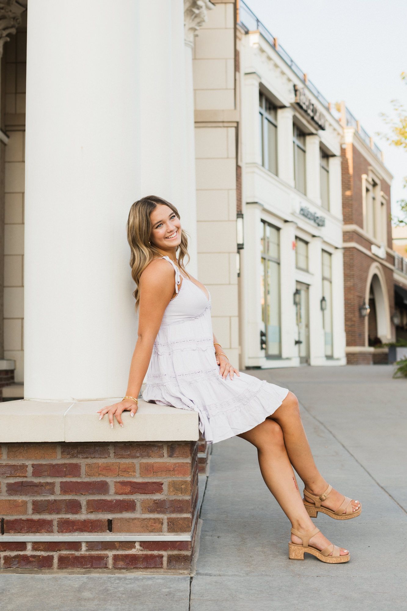 Young woman in a light sundress and wedge sandals sitting on a brick ledge, smiling outdoors on a city street.