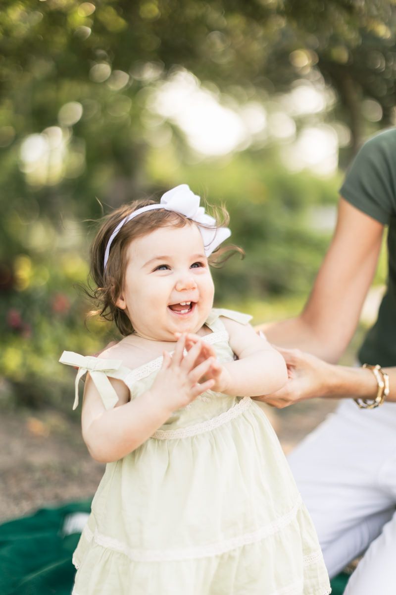 Baby in cream-colored dress laughing joyfully outdoors.