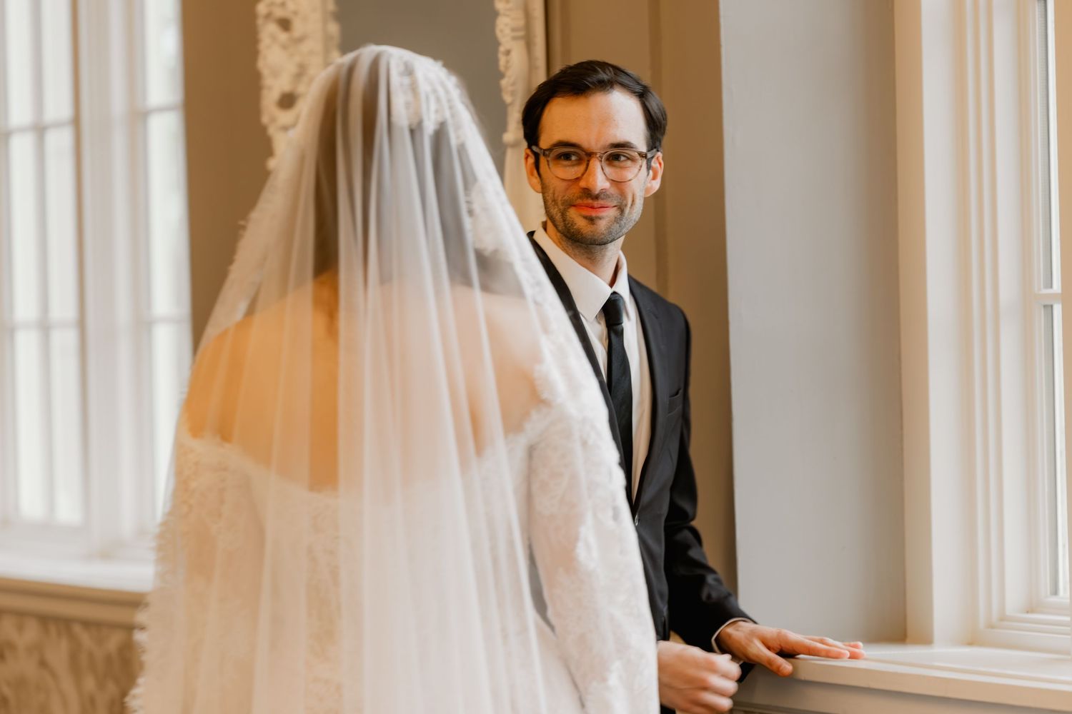 Groom in black suit smiling at bride in white wedding dress and veil during indoor wedding ceremony.