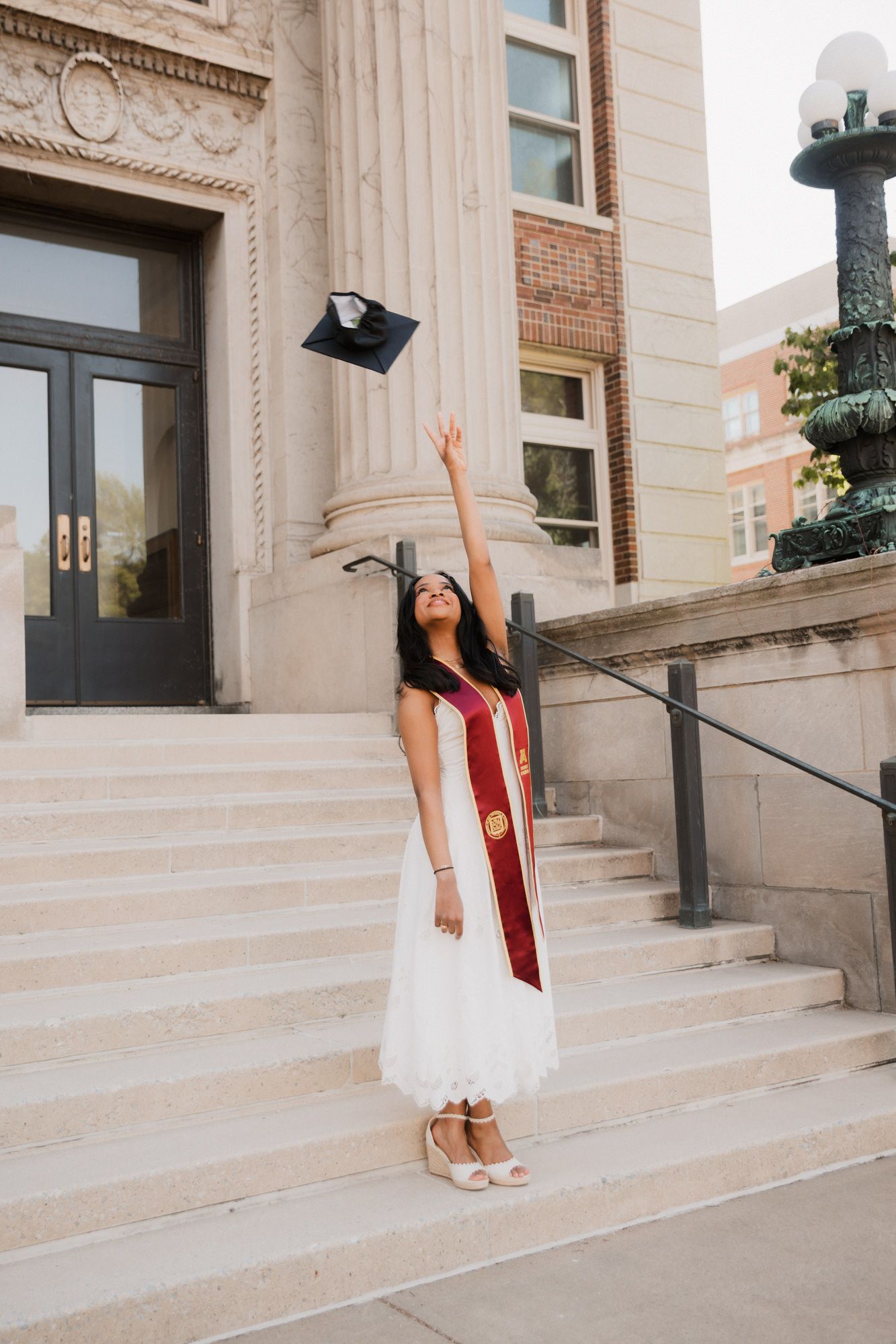 A graduate in a white dress and red sash tosses her cap in the air on university steps.