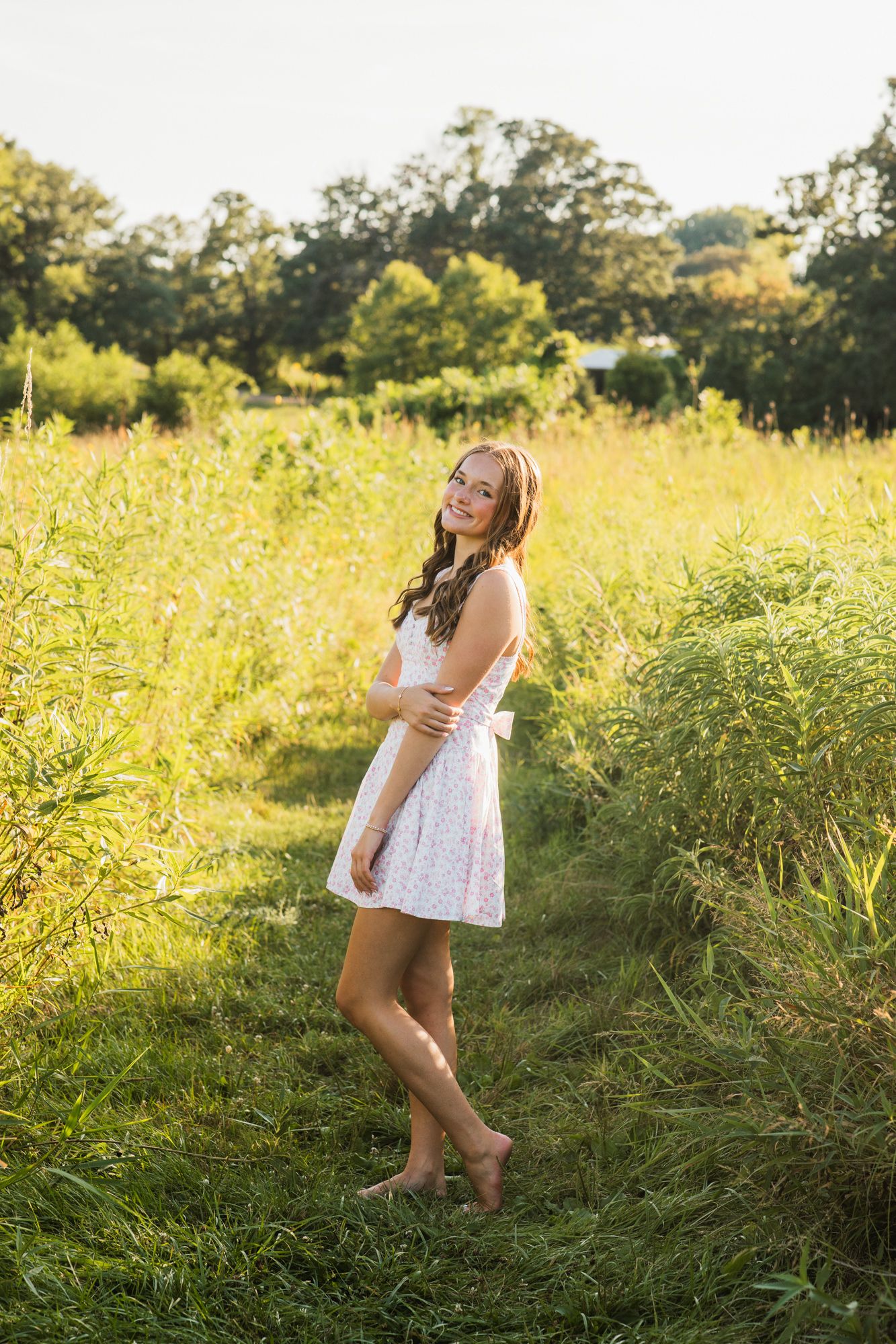 Young woman in white sundress standing barefoot in lush green field during golden hour sunlight.