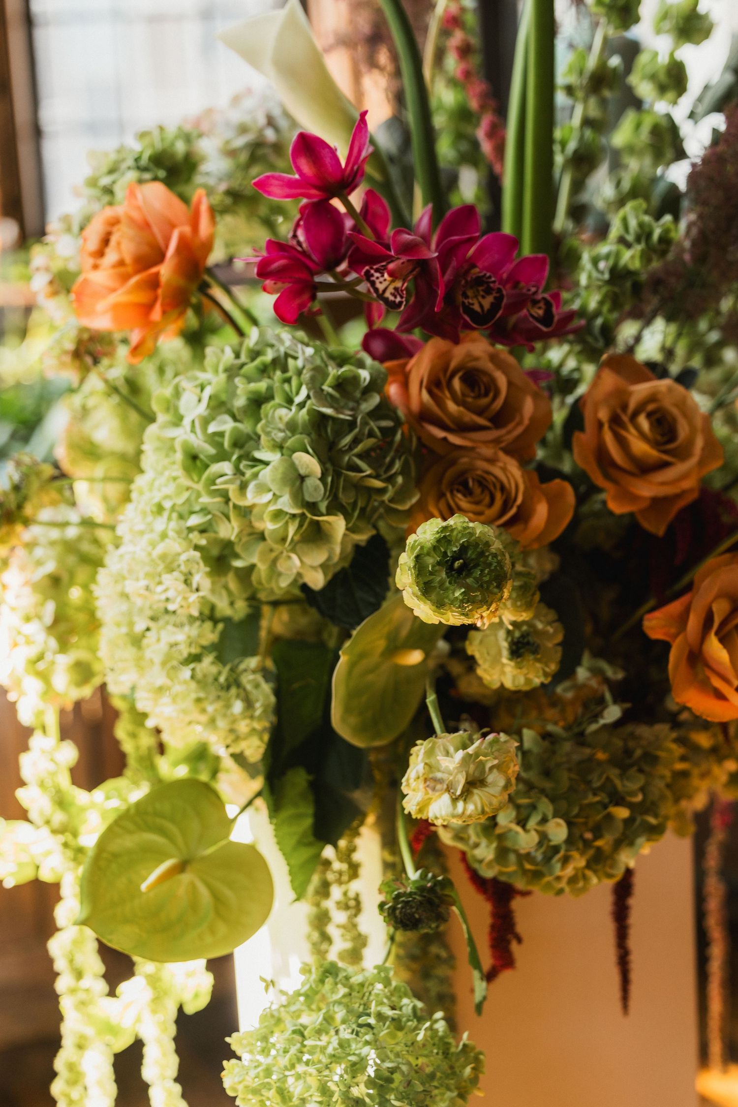 Lush floral arrangement with orange roses, magenta orchids, green hydrangeas, and anthurium in a rich autumn palette.