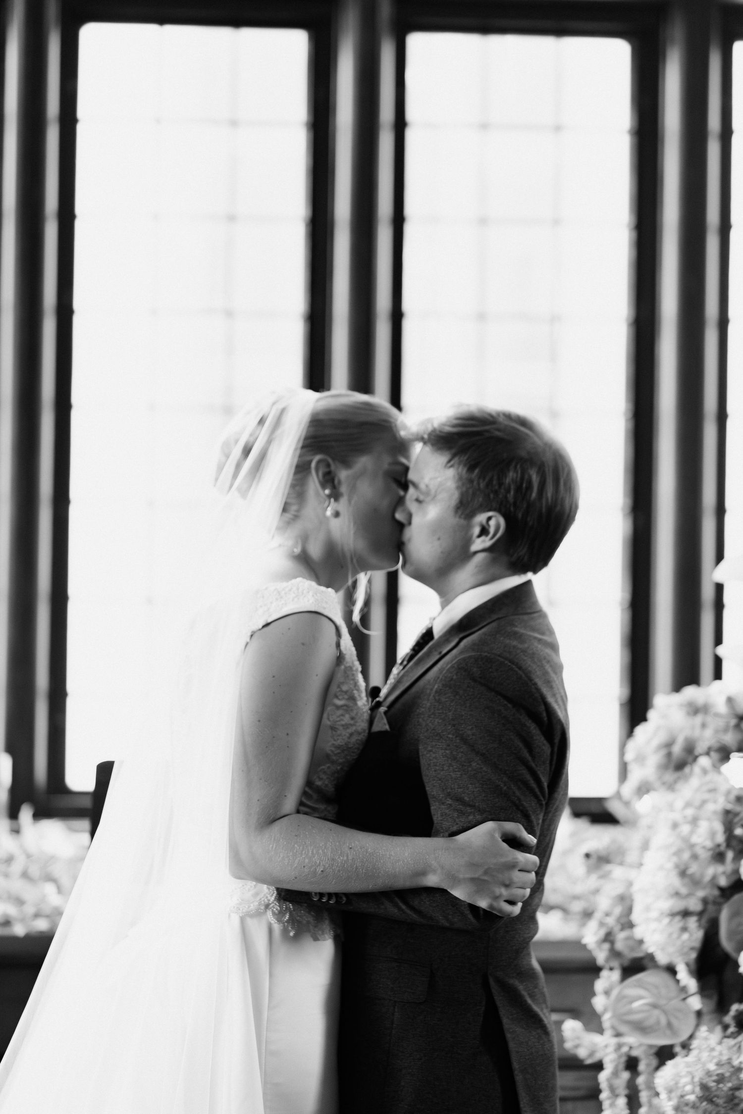 Black and white photo of bride and groom sharing their first kiss during wedding ceremony near tall windows.