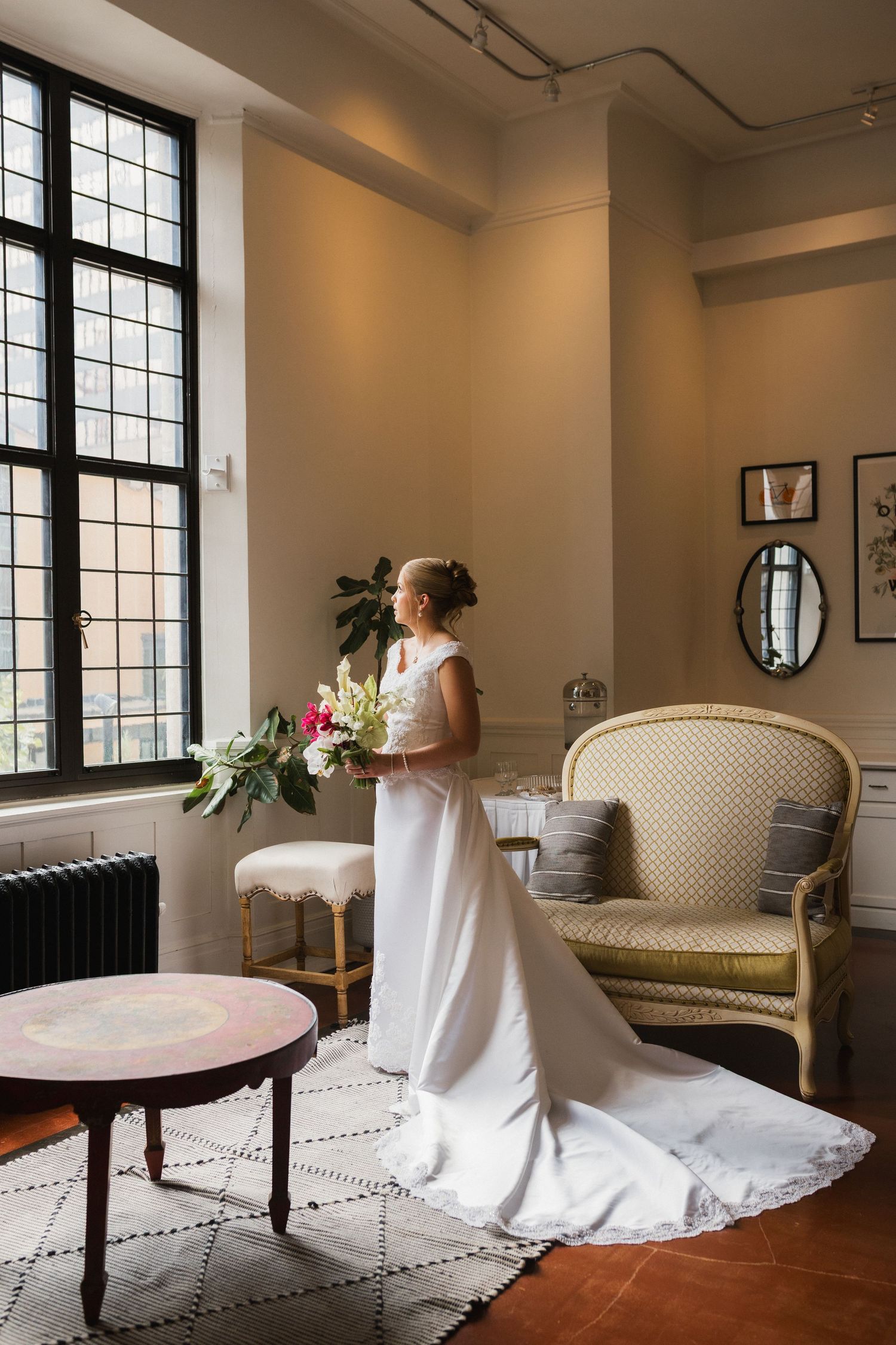 Bride in white gown holding floral bouquet stands by large window in elegant room with vintage furniture.