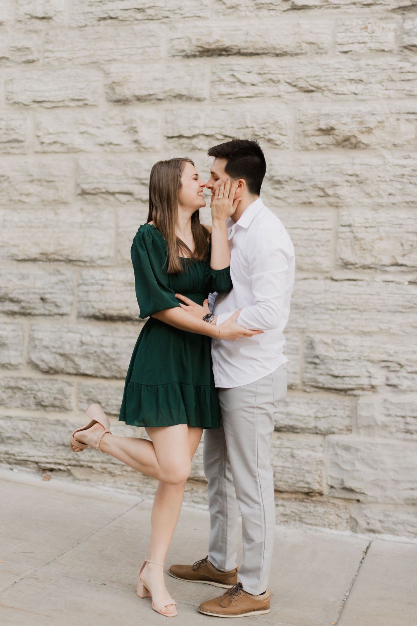 Happy couple laughing and embracing in front of a stone wall during an outdoor engagement photo session.