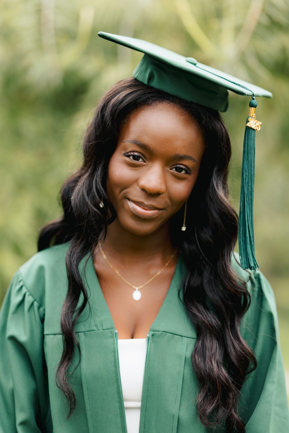 Graduate in green cap and gown with tassel wears delicate jewelry against garden backdrop.