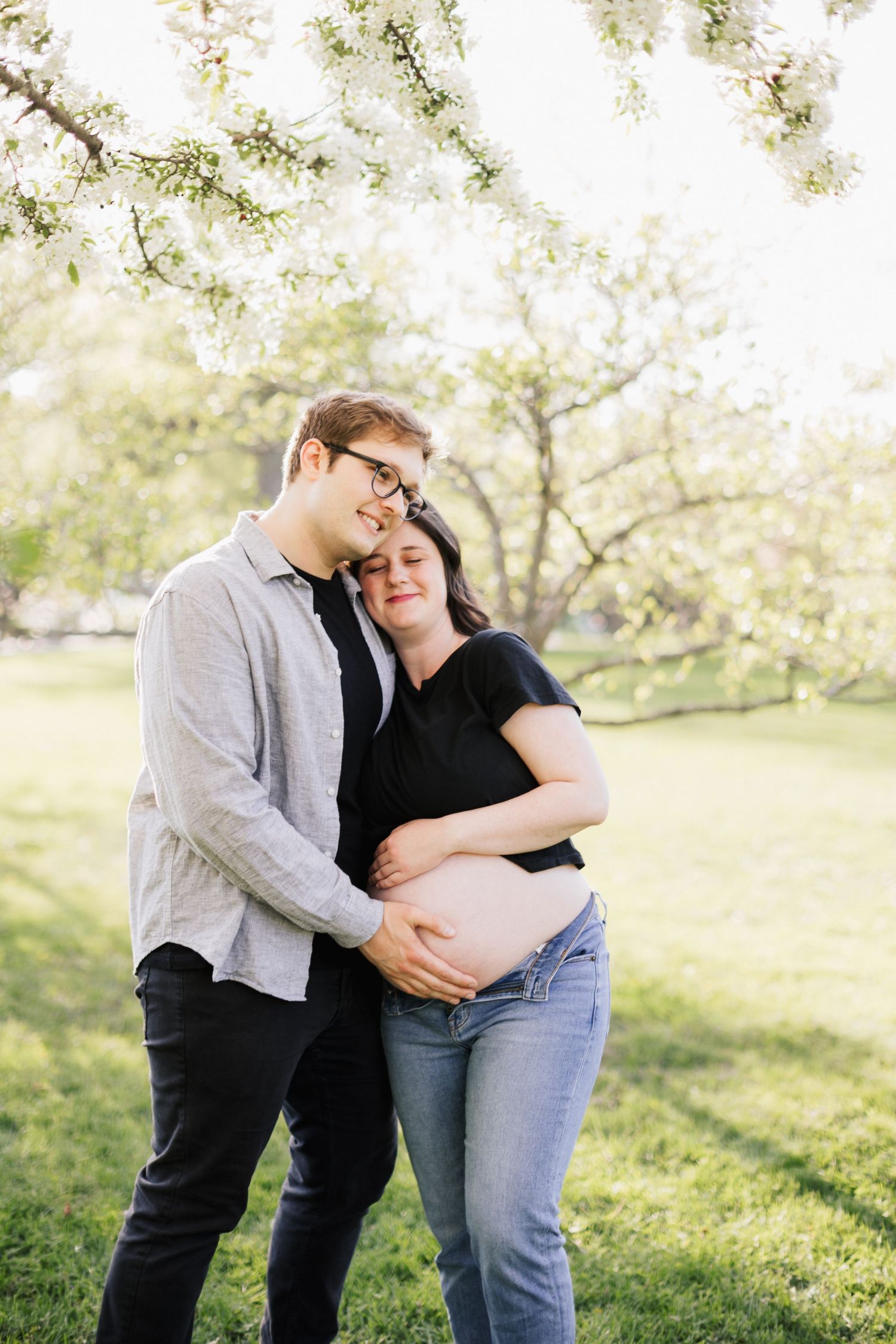 Expecting couple embraces under blooming spring trees.