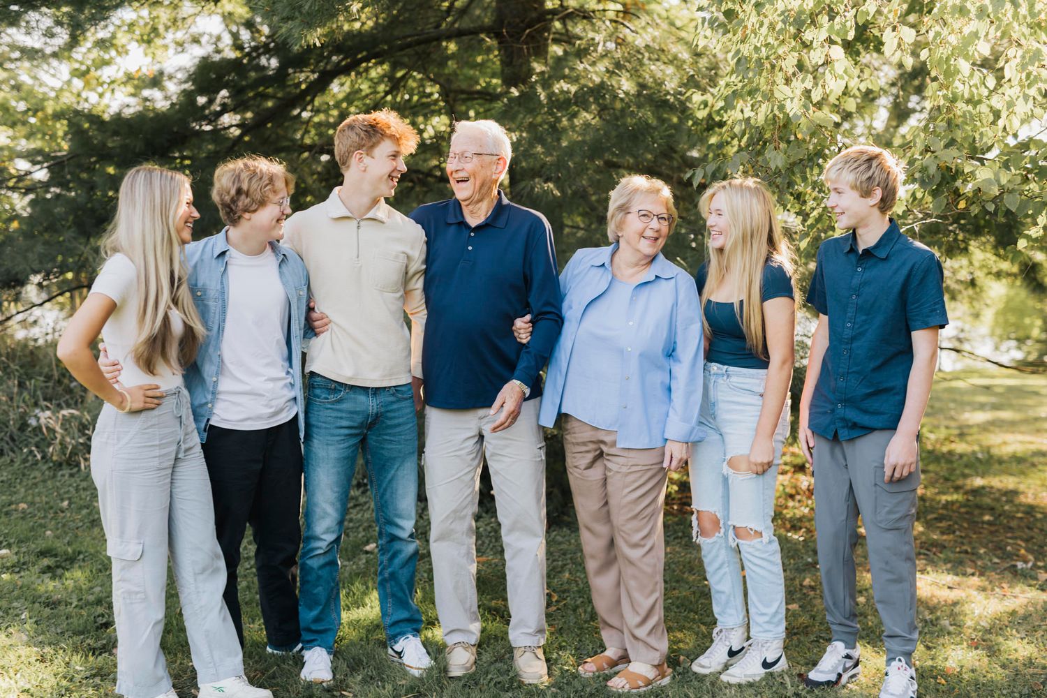 Extended family members line up casually for group photo in sunlit park setting.