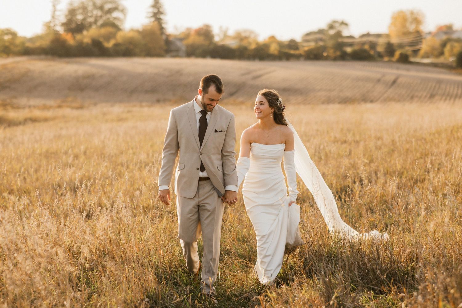 Bride in a white strapless gown and groom in a tan suit walk hand in hand through a golden grass field at sunset.