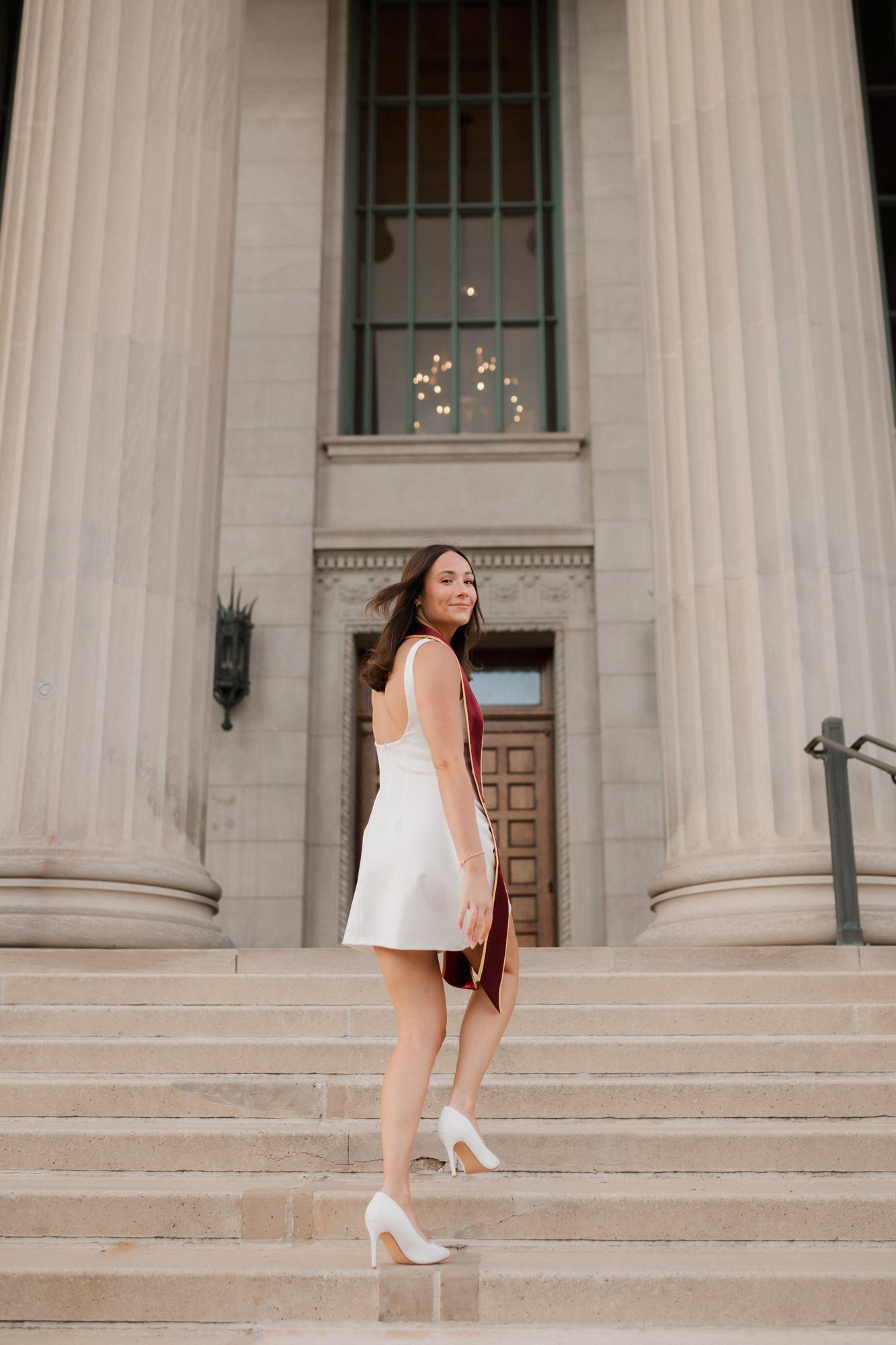 College graduate in white dress and maroon stole poses on steps of a neoclassical building.