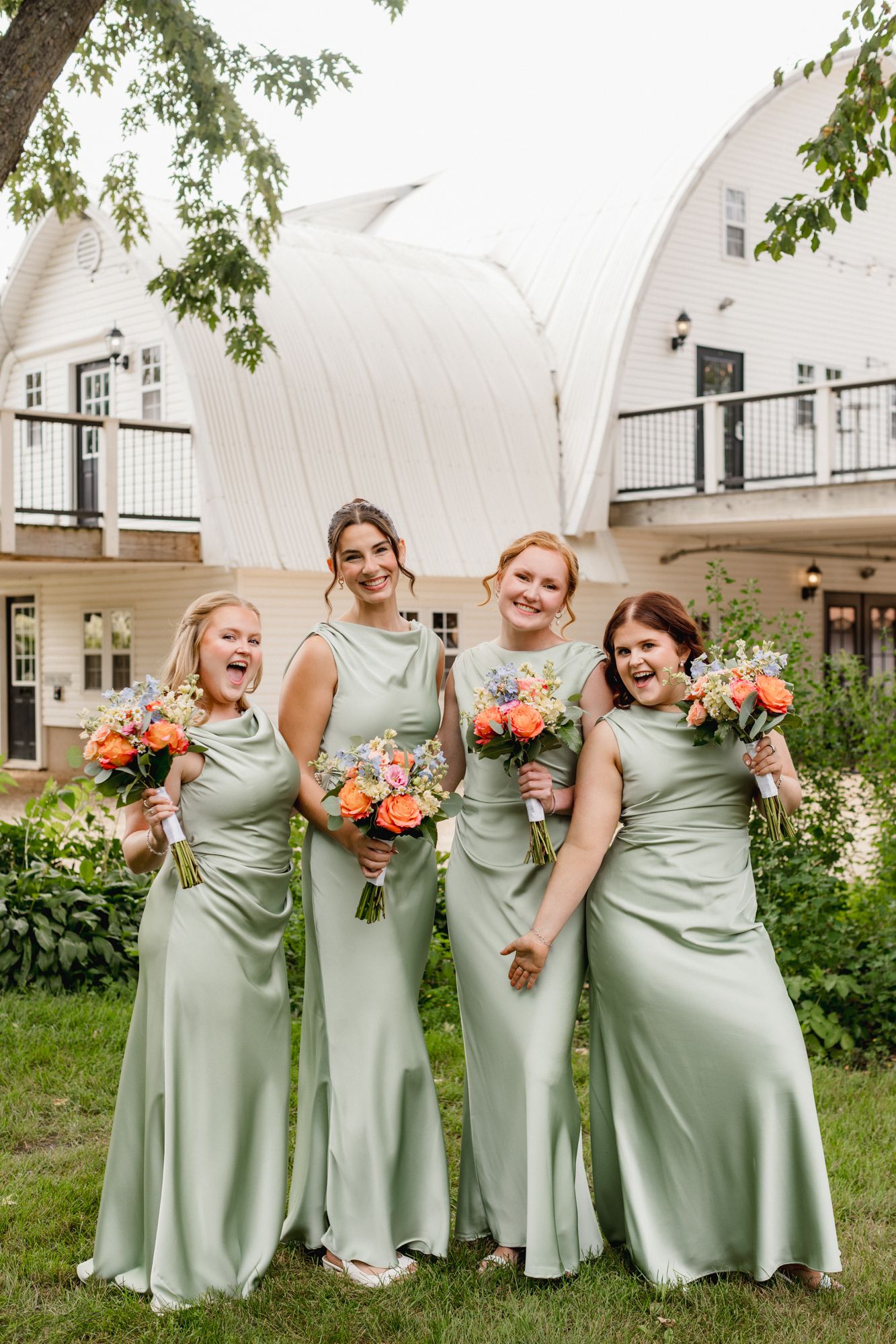 Four bridesmaids in sage green satin gowns holding colorful bouquets laugh together outdoors at a wedding venue.