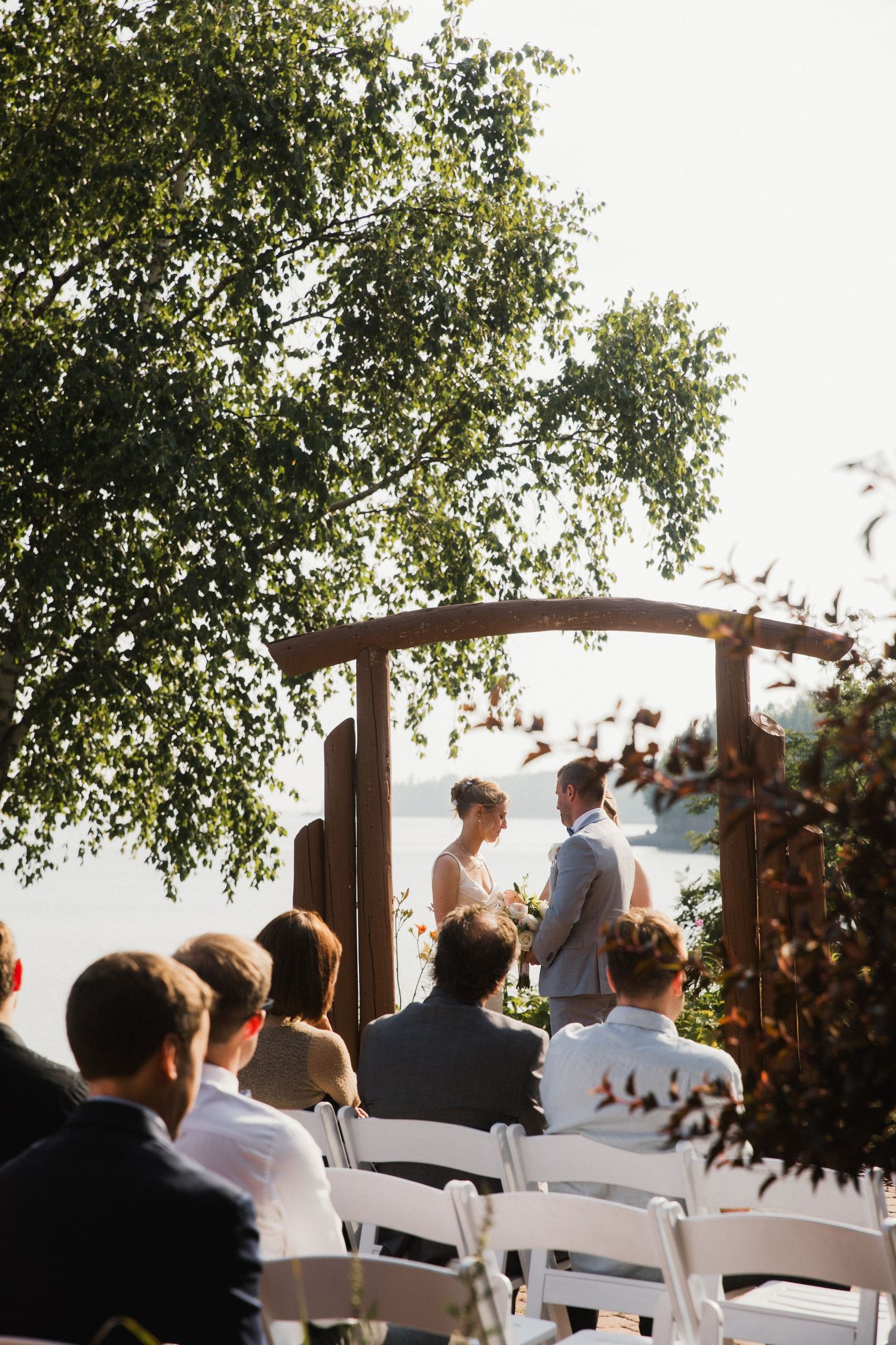 Outdoor wedding ceremony with couple at wooden arch altar, guests seated in white chairs, surrounded by lush green trees.