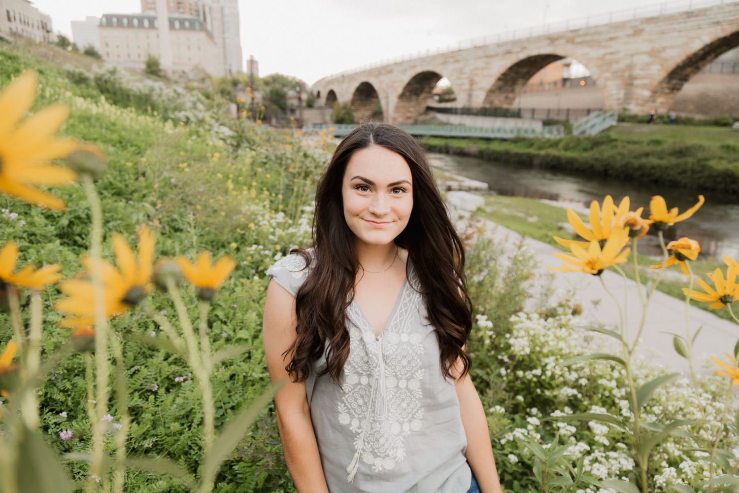 Young woman with dark hair smiling among yellow wildflowers with a stone bridge and river in the background.