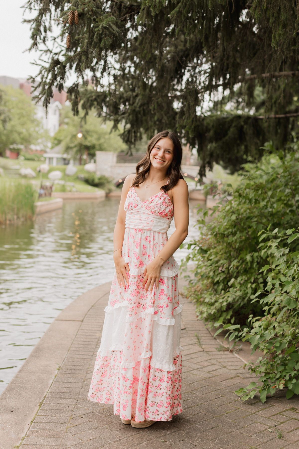 Smiling woman in pink floral maxi dress standing by a waterway surrounded by lush greenery.