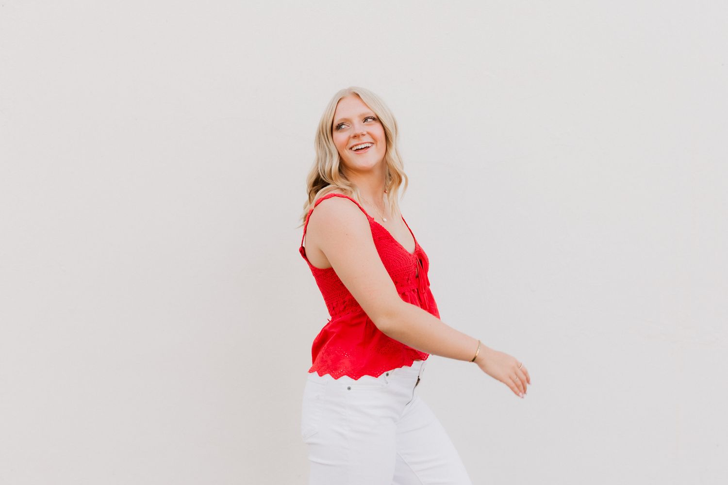 Smiling blonde woman in red sleeveless top and white jeans posing against a light gray background.