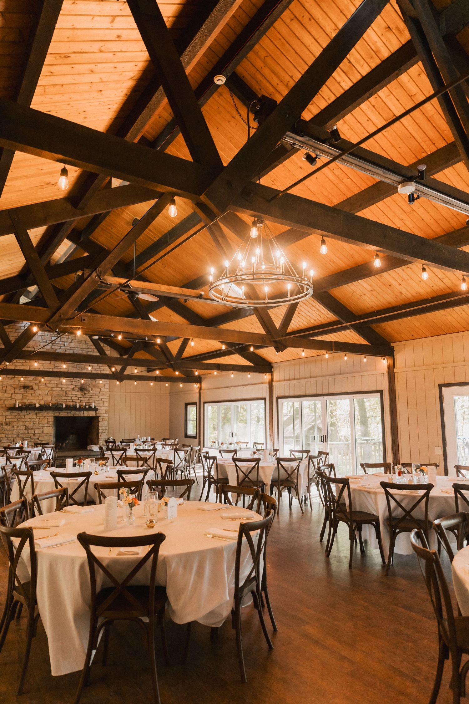 Rustic wedding reception venue with exposed wooden beams, white walls, and round tables set for dining under string lights.