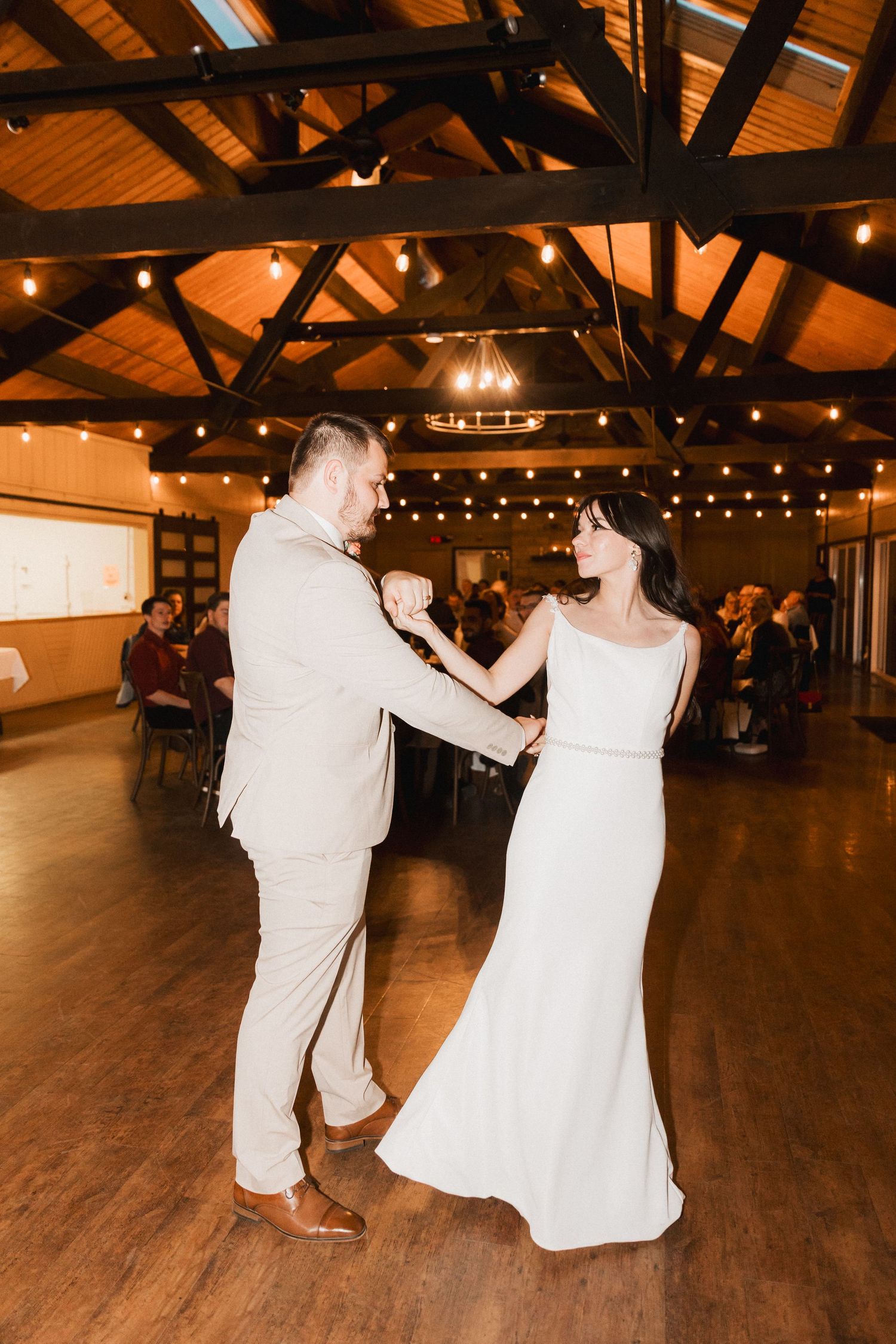 A couple dances together in a rustic venue with wooden beams and string lights creating a warm atmosphere.