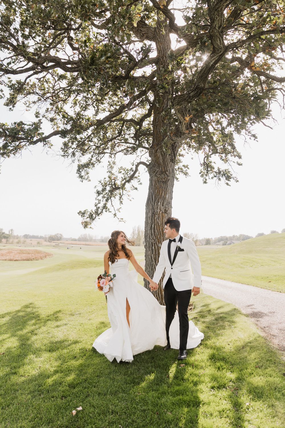 A couple in wedding attire shares a romantic moment under a large tree on a grassy hillside during sunset.