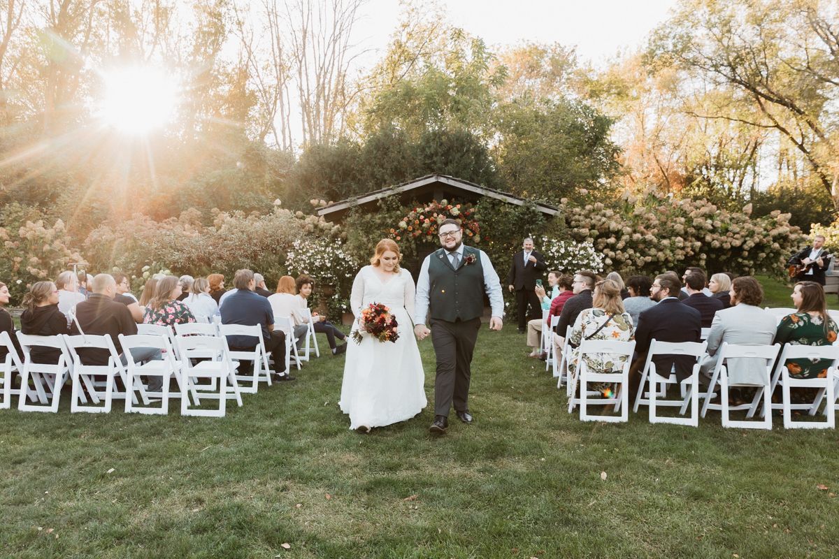 Couple walks down aisle at sunset during outdoor garden wedding with white chairs.