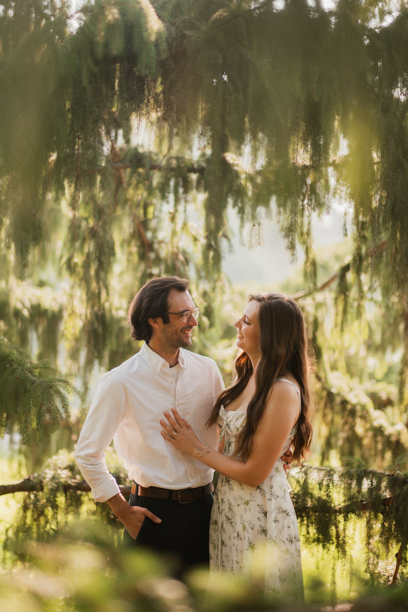 Couple sharing a romantic moment under weeping willow tree in golden hour light, woman in floral dress, man in white shirt.