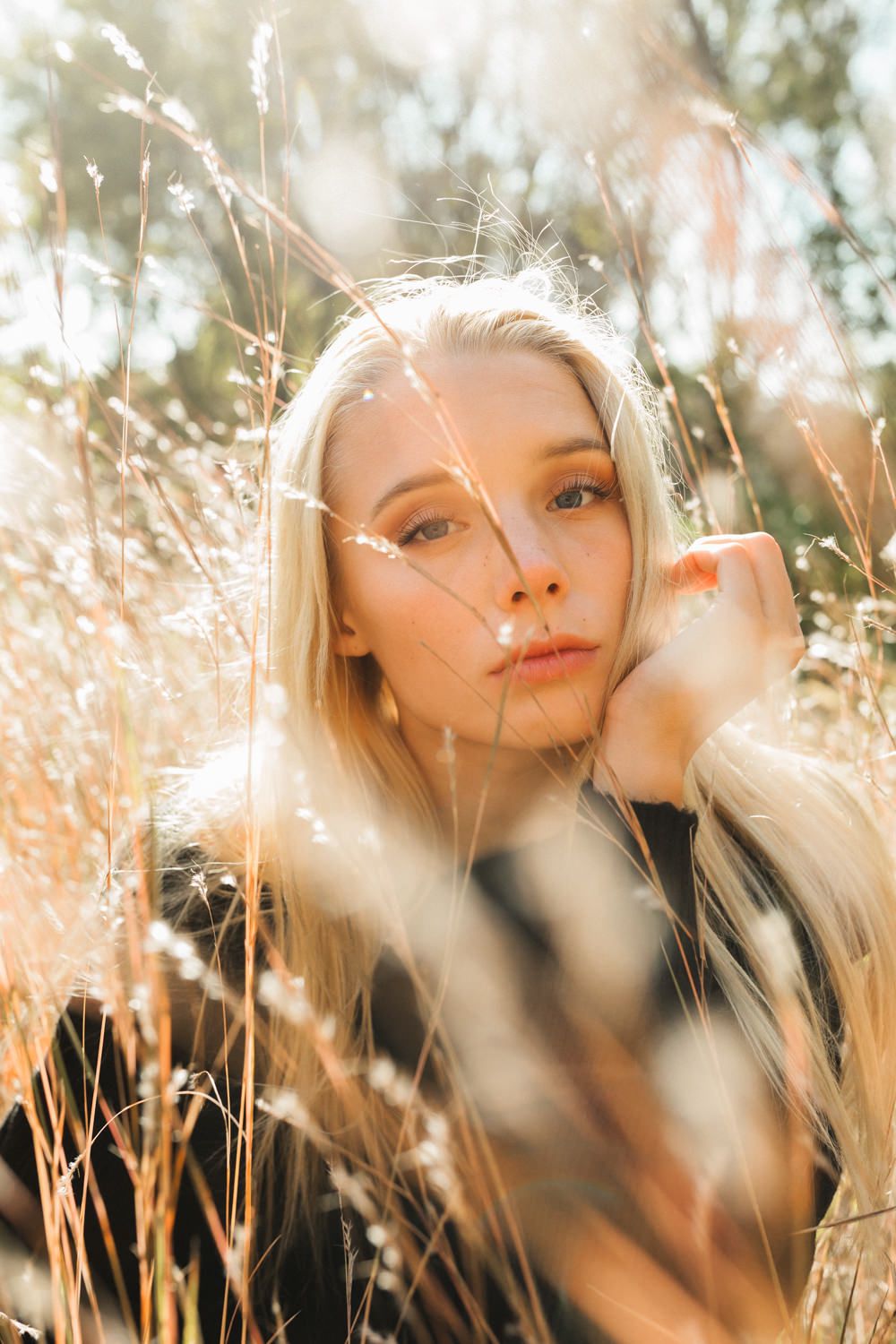 Blonde woman sitting in golden sunlit tall grass field, resting chin on hand, with a dreamy bokeh outdoor background.