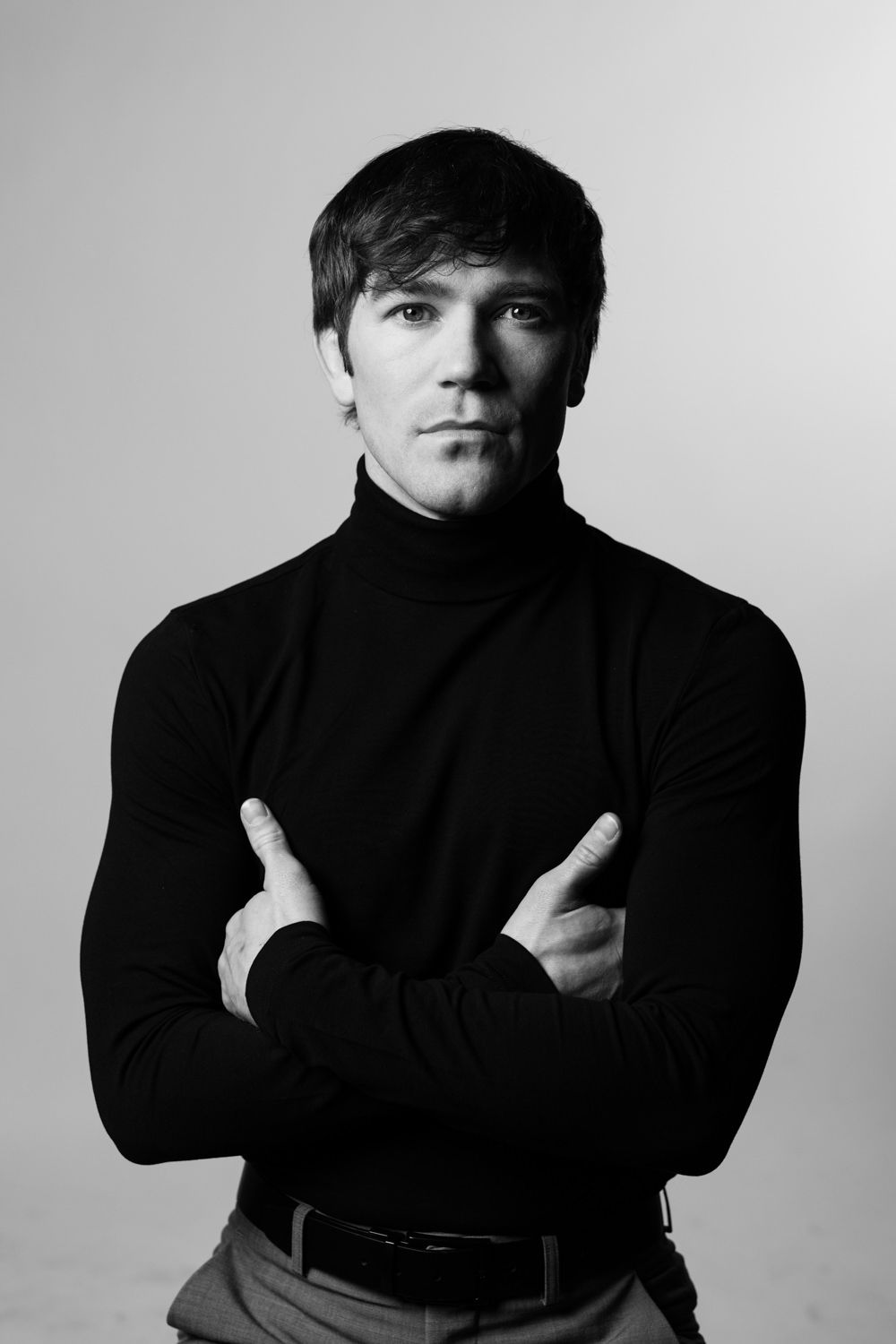 Black and white portrait of a serious man in a black turtleneck sweater with arms crossed against a light background.