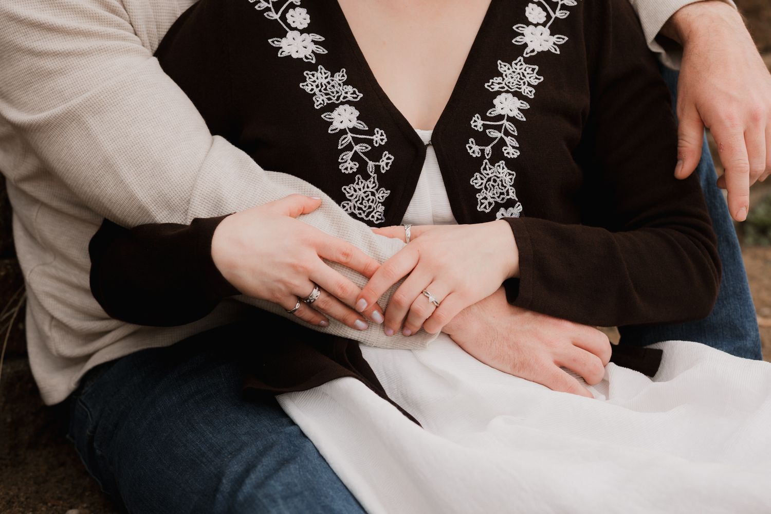 Couple sitting closely together, hands intertwined, wearing embroidered black top and jeans outdoors.