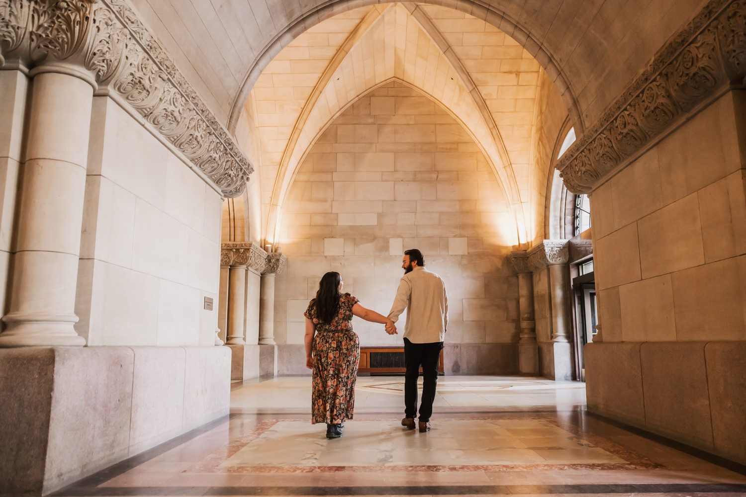 Two people walk hand in hand down an elegant stone corridor with vaulted ceilings and arched architecture.