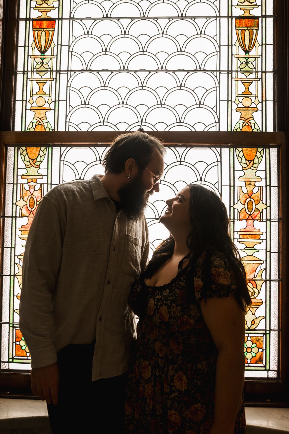 A romantic silhouette of a couple sharing a tender moment in front of an ornate stained glass window with geometric patterns.